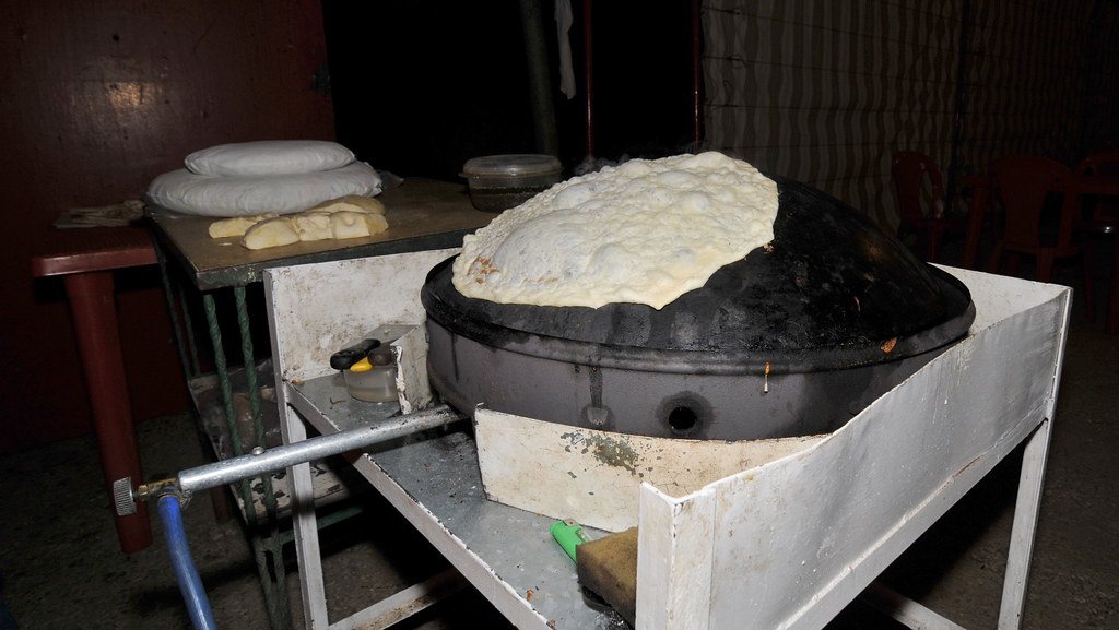 A thin sheet of markouk bread lying over a black domed saj heated by a gas burner, with dough balls on the bench behind