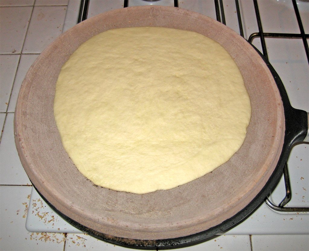 A round of pale raw piadina dough on a round terracotta testo set over a gas hob