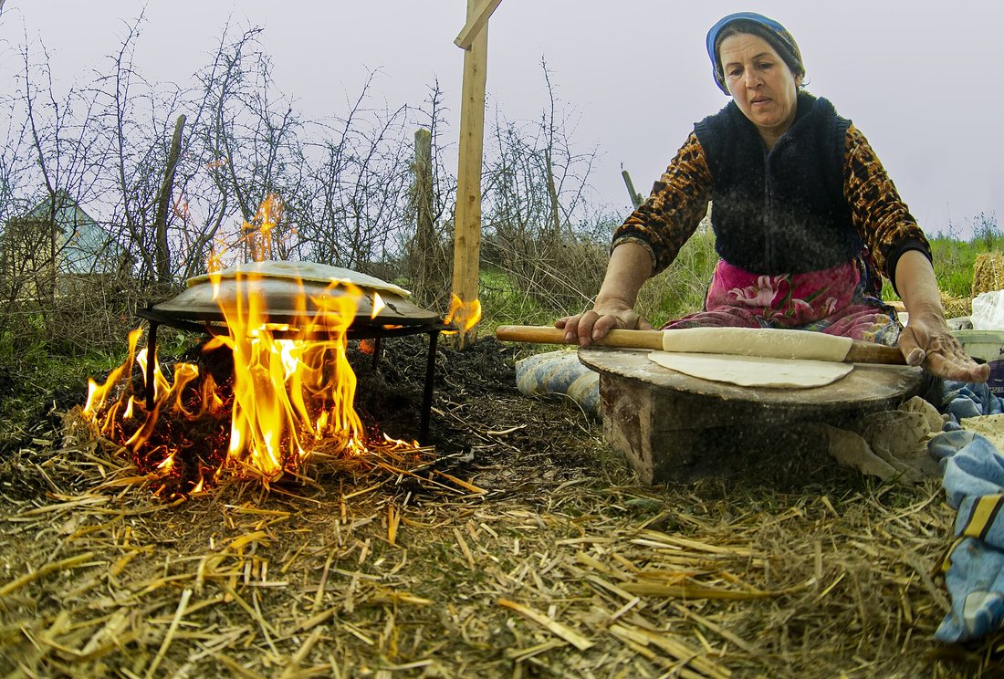 A woman outdoors rolling a thin sheet of dough with a long pin beside a griddle set over an open wood fire