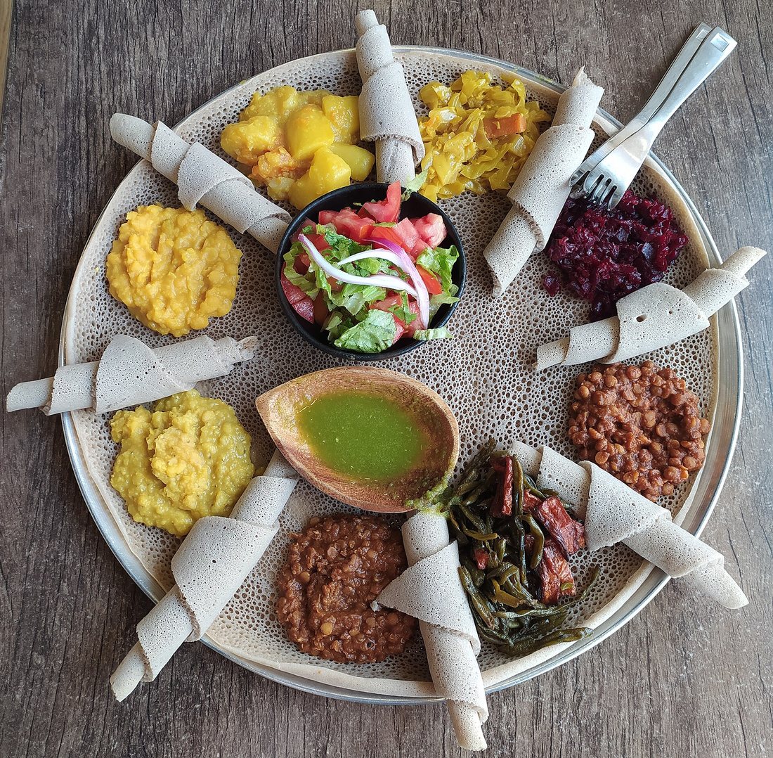 Top view of a round platter of injera covered with several vegetable stews and a salad, with rolled injera arranged around the edge