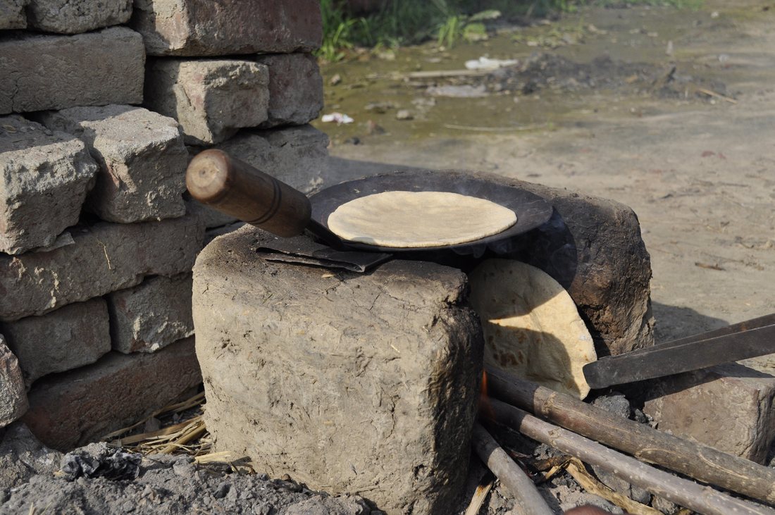 A roti cooking on an iron tava over a mud-and-brick stove fired with sticks, outdoors