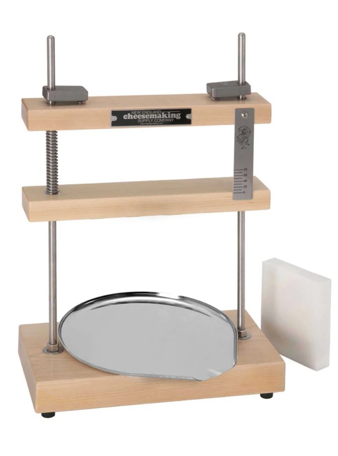 The screw press: two stainless guide rods, a fixed top bar, a coil spring around the central screw, and a
calibrated scale plate on the right that reads the spring's compression. Stainless drip tray, white plastic
follower.