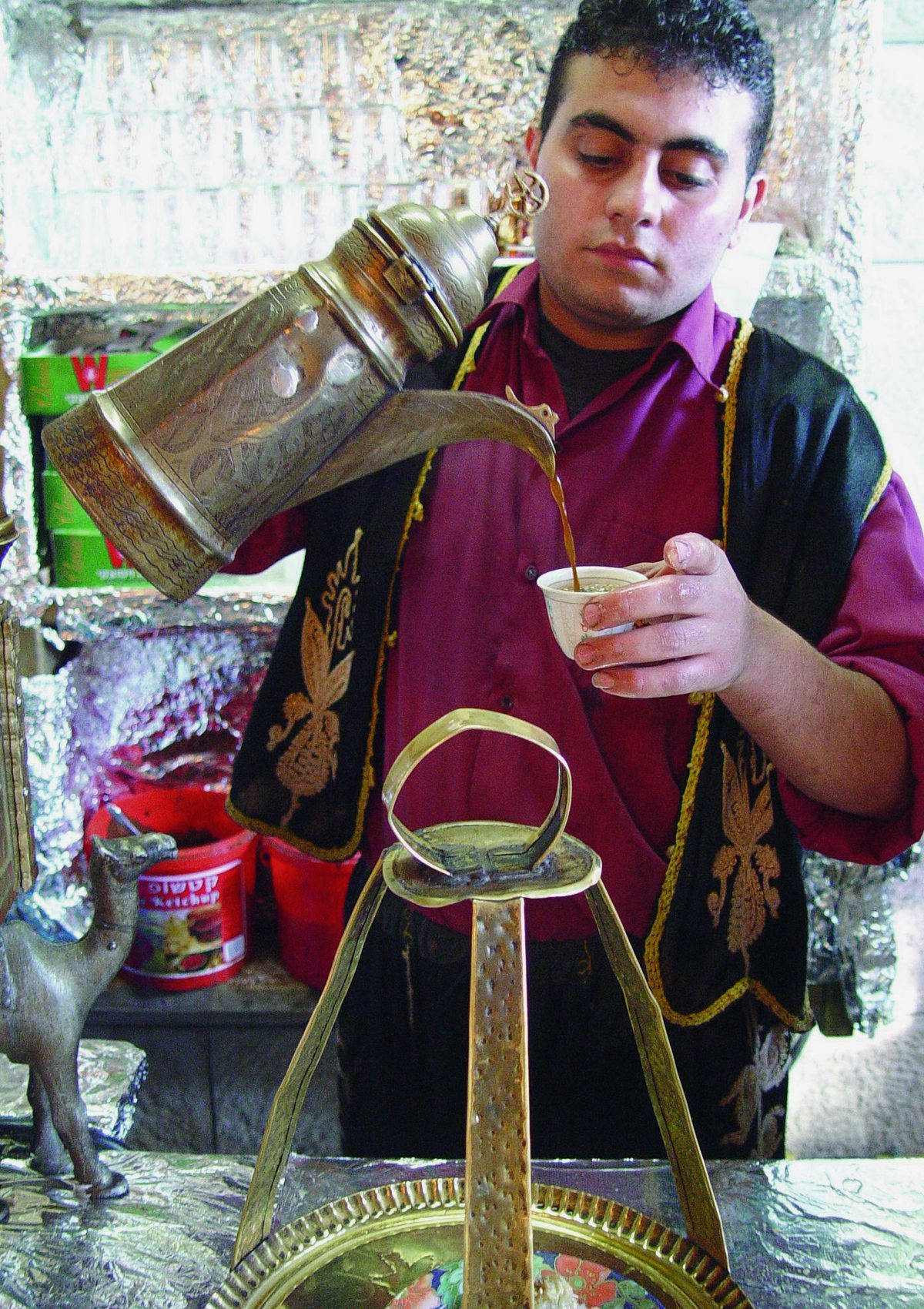 A street vendor pouring cardamom coffee from an ornate long-spouted pot into a tiny cup — the Gulf and Levantine qahwa tradition, spiced, poured from a height, served in a mouthful