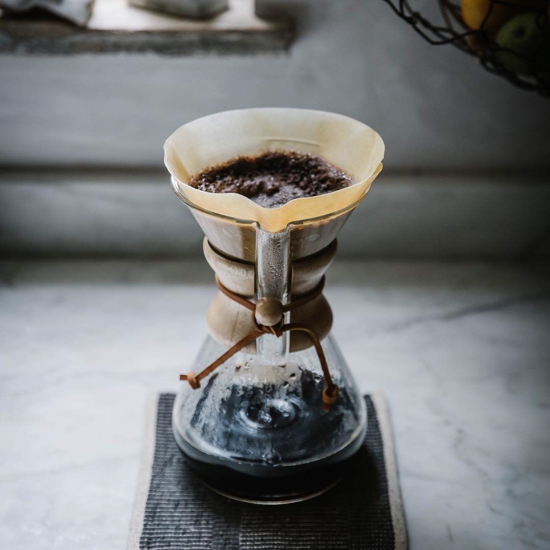 A Chemex mid-brew, the thick folded filter holding the grounds above the wood collar and leather tie