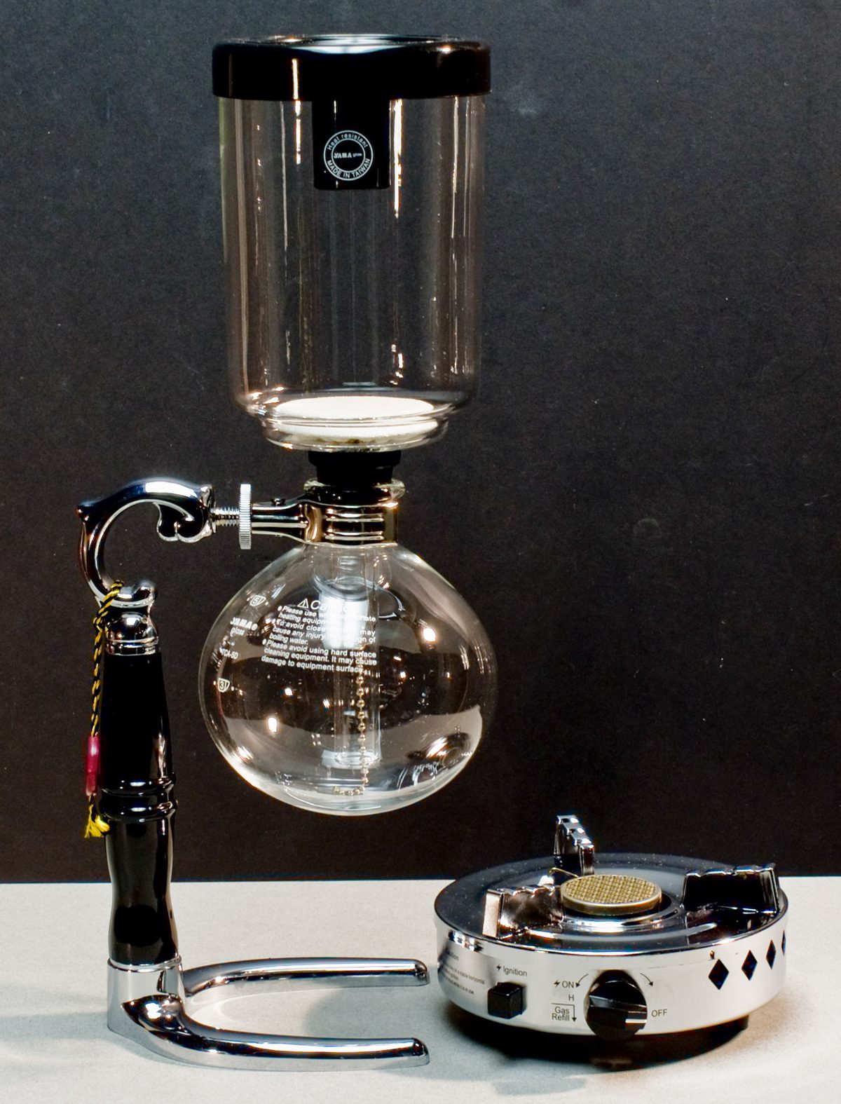 A glass siphon set up before the brew — upper globe with its filter seated, water in the lower bulb, butane burner standing by