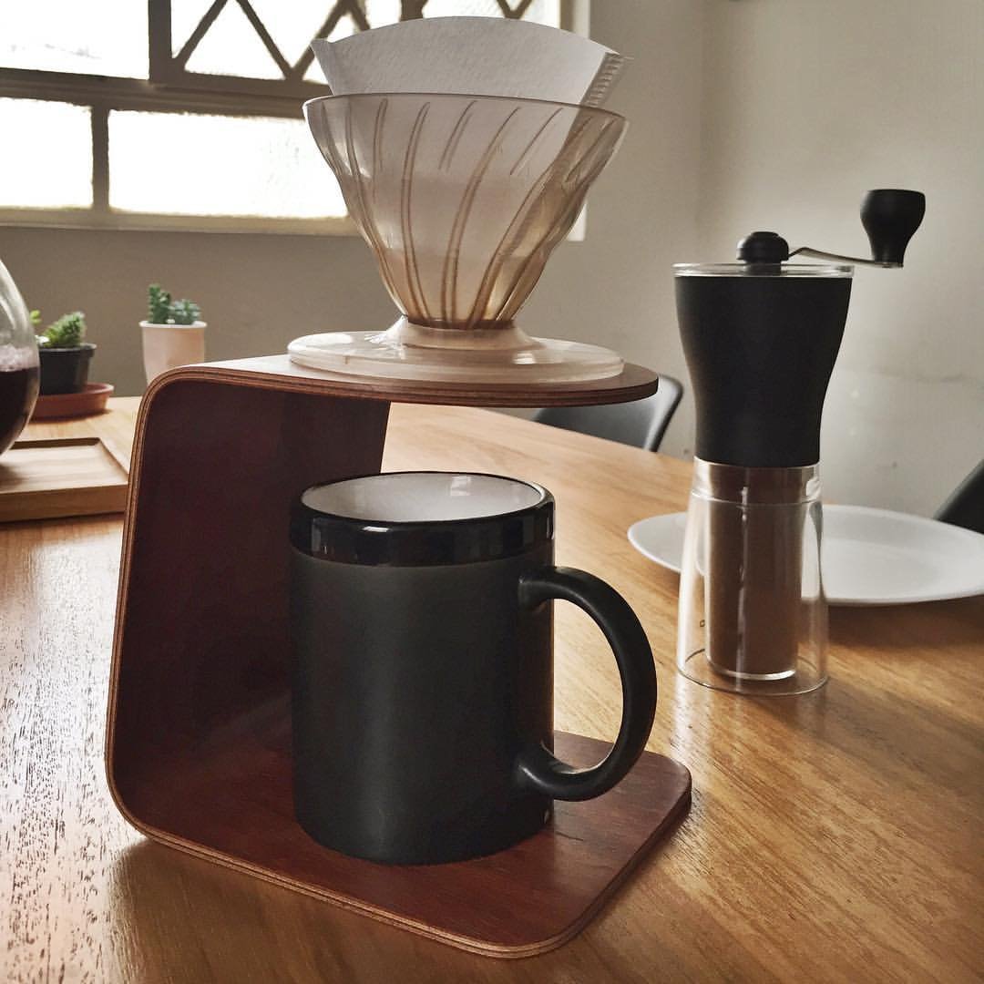 A paper-lined cone dripper on a bent plywood stand over a black mug, with a slim hand grinder beside it holding ground coffee in its clear base