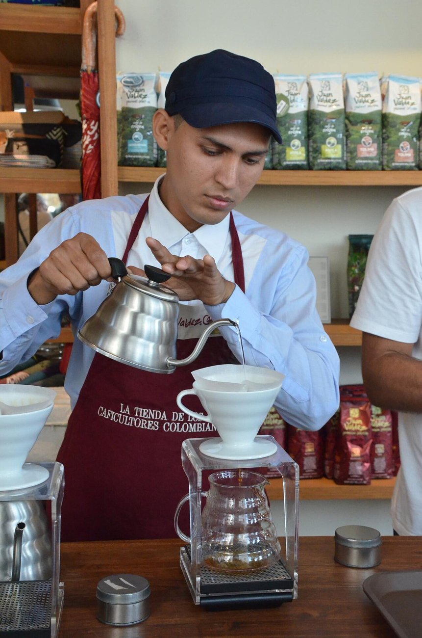 A barista in a maroon apron pours from a steel gooseneck kettle into a white cone dripper that sits on a glass server on a scale