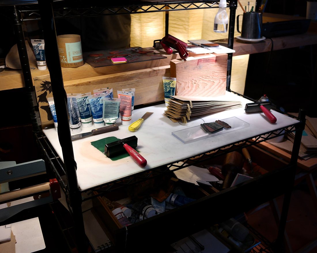 A workbench with ink rollers, a printing block and a stack of paper coffee bags