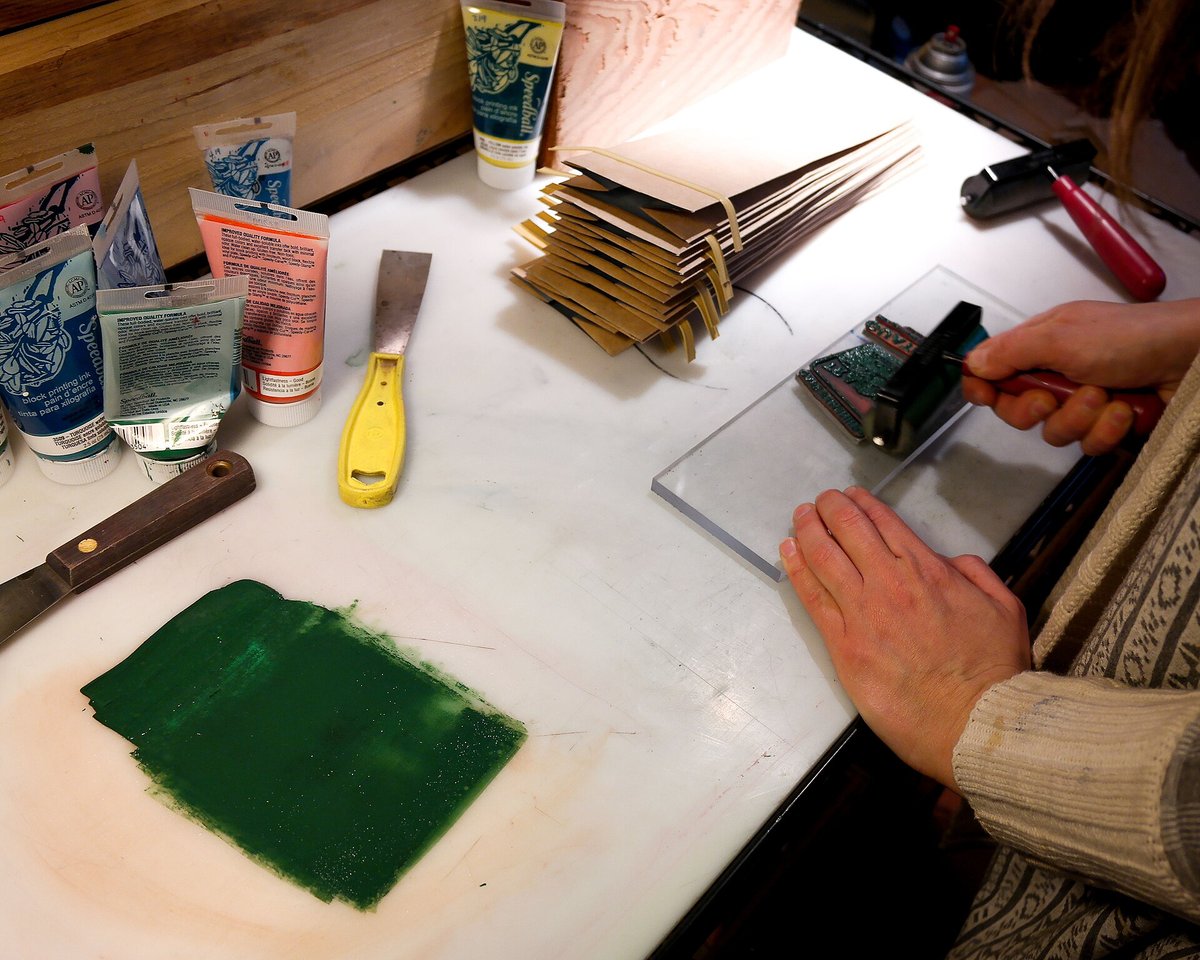 A hand inking a square printing block on a sheet of glass with a roller, beside a stack of plain kraft coffee bags and a slab of green ink
