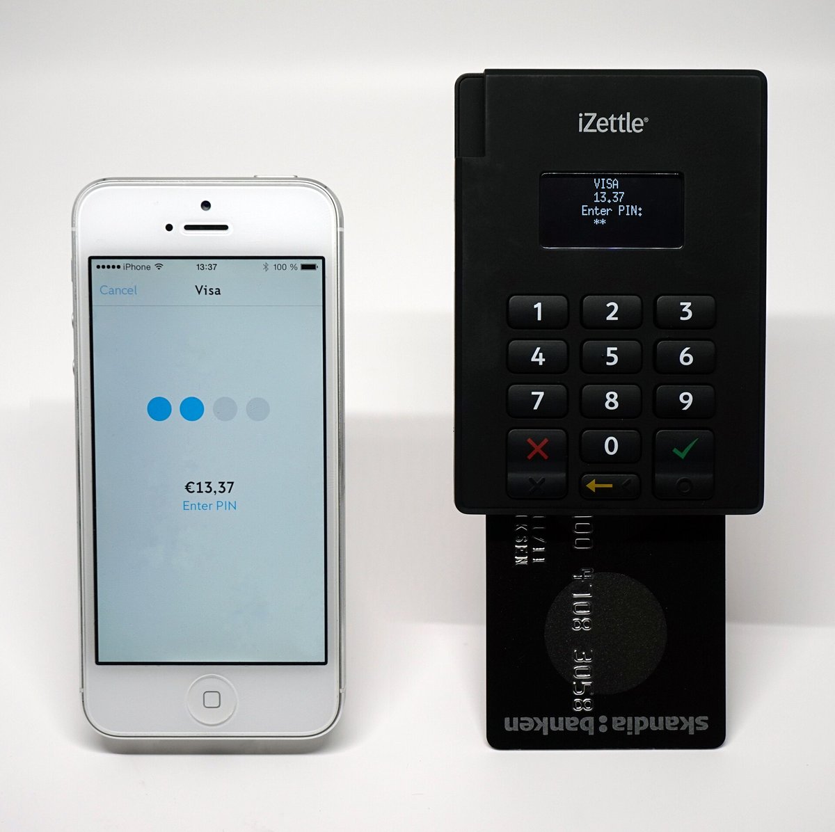 A small black card reader with a keypad, standing beside a smartphone showing a payment in progress