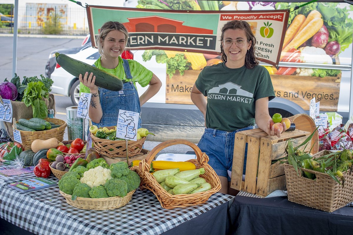 Two vendors smiling behind a farm stand of courgettes, broccoli, cauliflower and squash under a banner saying the market proudly accepts food-assistance benefits