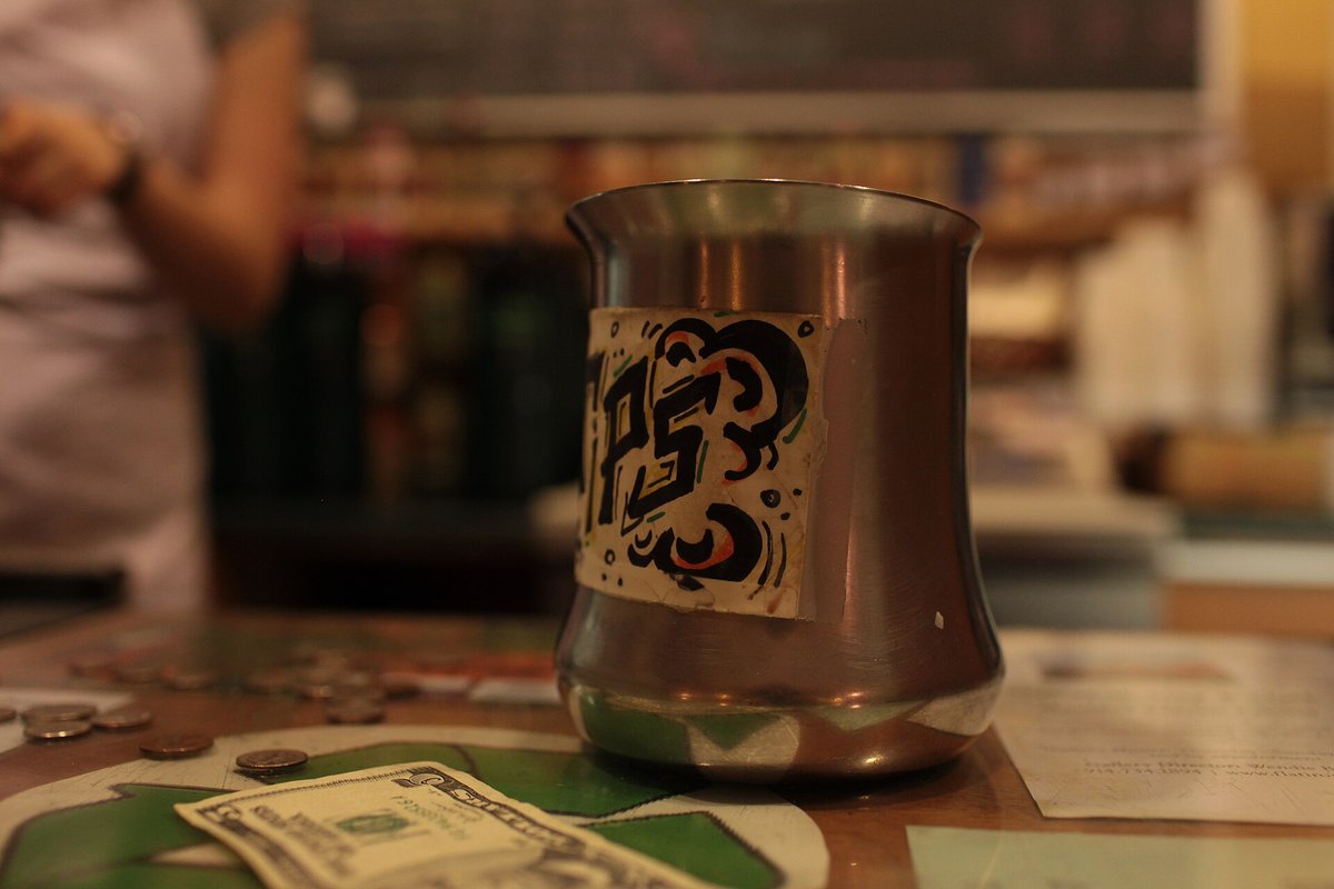 A metal jug with a sticker on it, used as a tip jar, with a dollar bill beside it on a counter