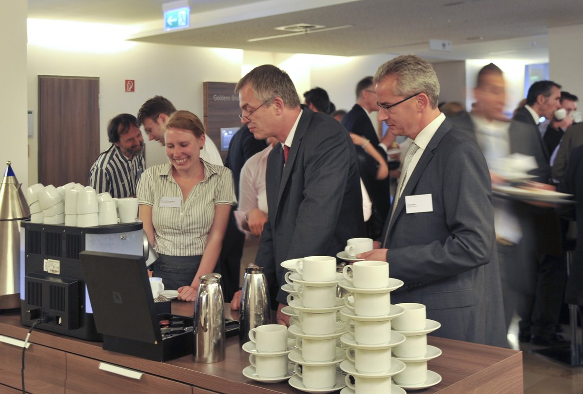 A long service counter at a conference with rows of white cups and saucers, insulated coffee pots and guests helping themselves