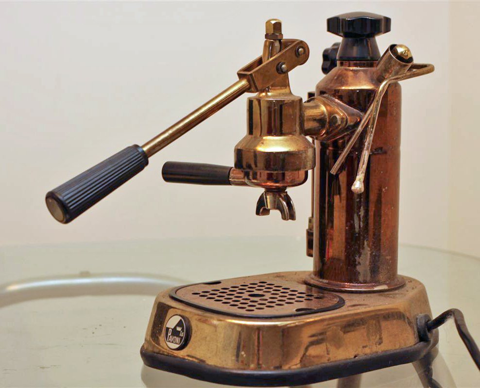 A small copper-and-brass lever espresso machine with a black-handled lever and portafilter on a round base