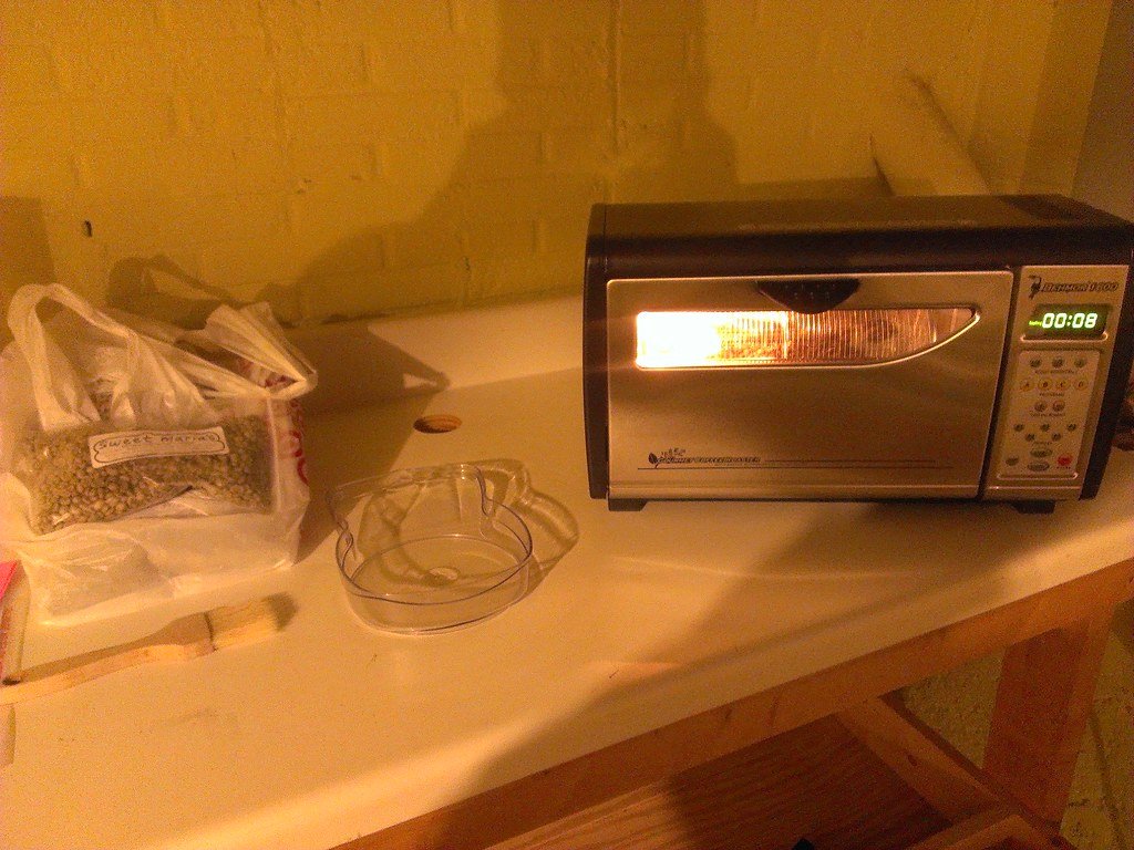A stainless Behmor drum roaster on a workbench, its window glowing orange, the timer reading 00:08, with a bag of green coffee and a clear chaff tray beside it