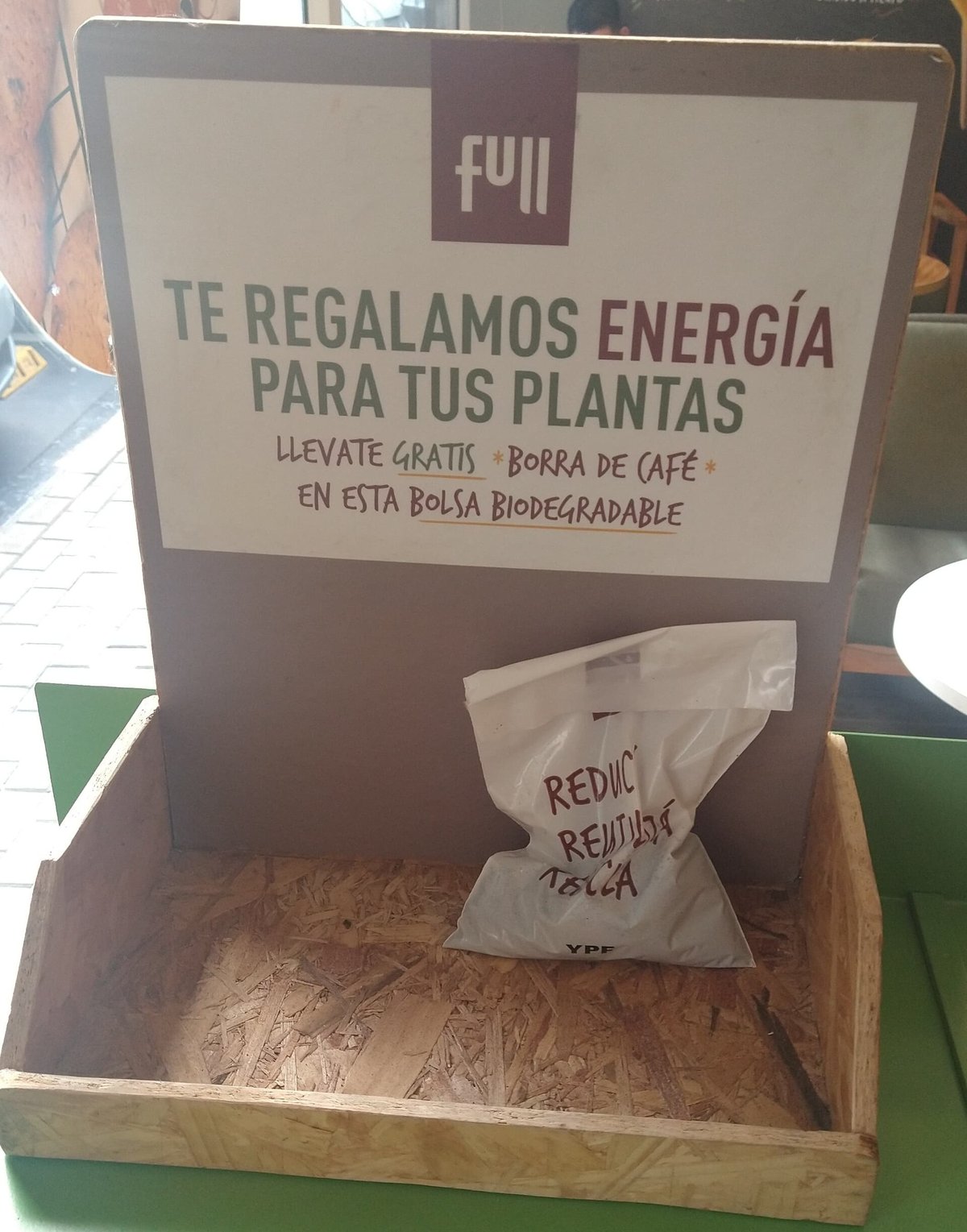 A cardboard counter display holding small clear compostable bags of used coffee grounds, with a sign in Spanish: we give you energy for your plants, take free coffee grounds in this biodegradable bag