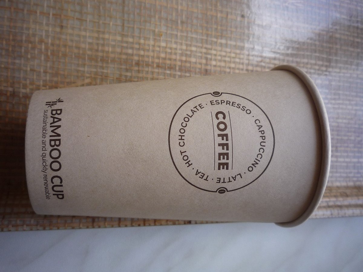 A kraft paper takeaway cup lying on its side on a woven mat, printed with a stamp that reads coffee, cappuccino, latte, espresso, tea, hot chocolate and, along the side, Bamboo Cup