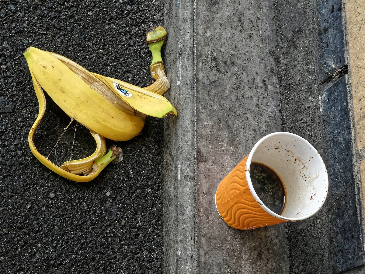 A disposable cup with an orange ribbed sleeve lying on its side on dark pavement next to a discarded banana skin, coffee dregs inside