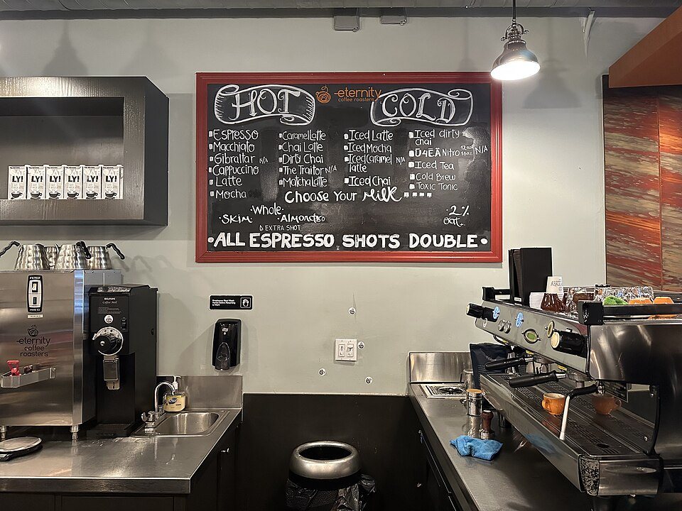 A chalkboard café menu in two columns headed Hot and Cold, above an espresso machine and grinder