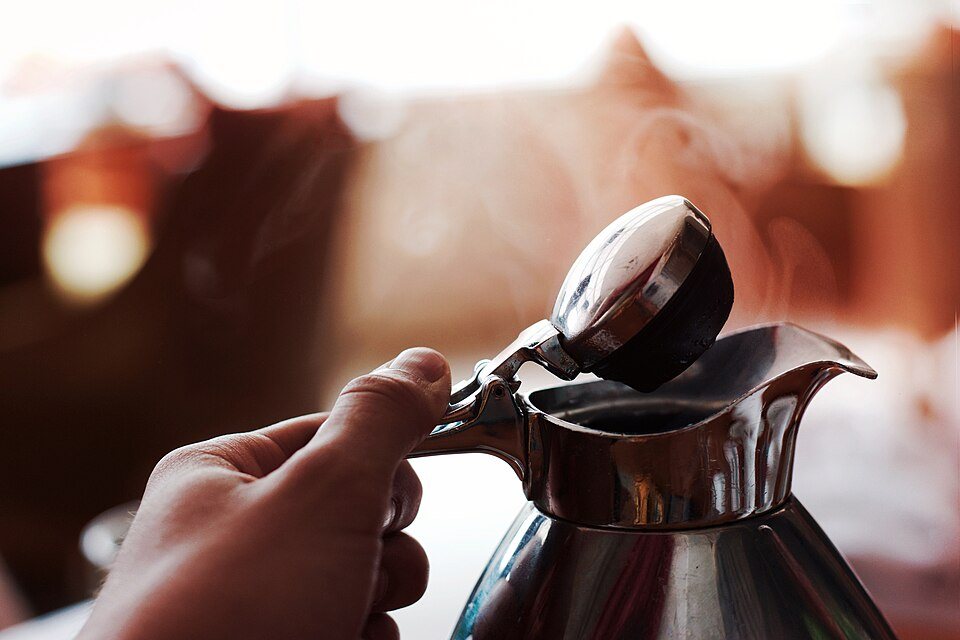 A hand lifts the hinged lid of a dark thermal coffee pot