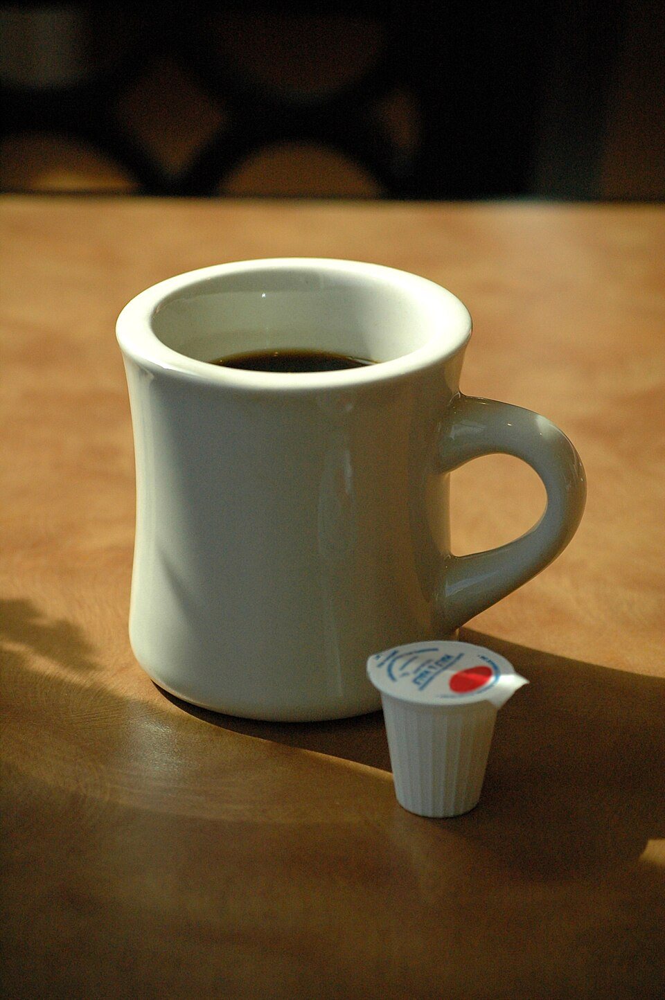 A thick white diner mug of black coffee on a wooden table beside a creamer cup