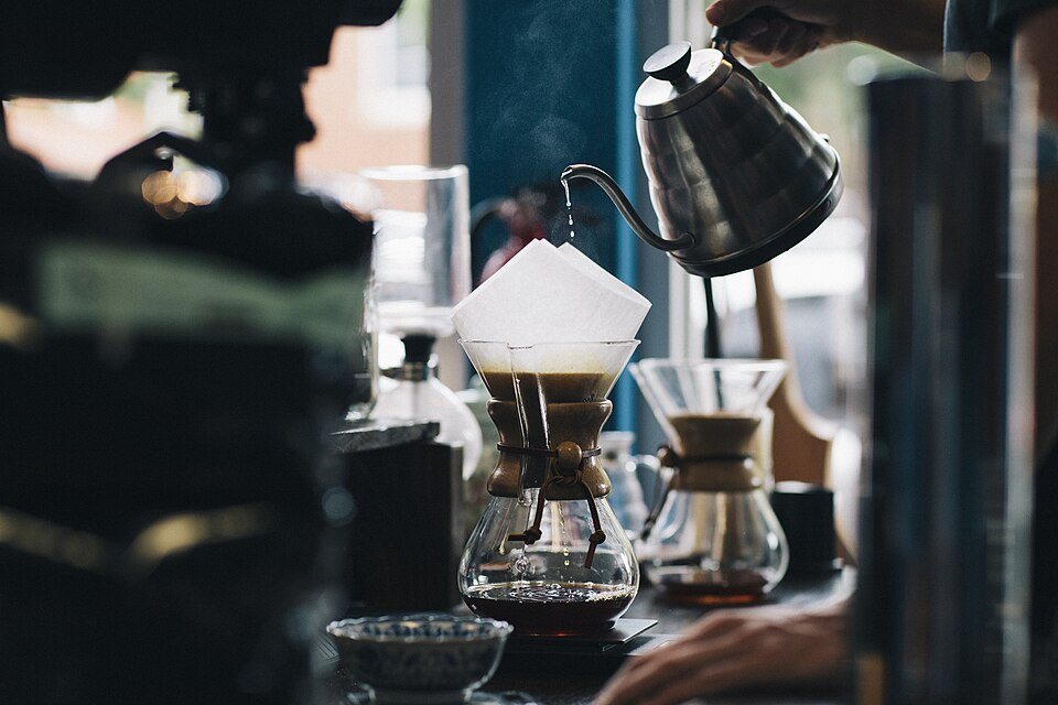 A gooseneck kettle pouring into a paper filter in a glass Chemex, in a dim café