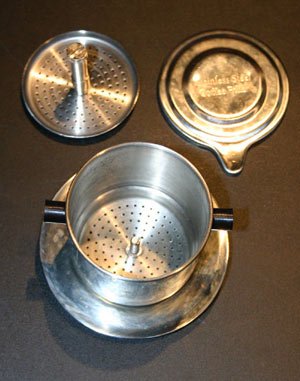 A Vietnamese phin taken apart on a dark table: the perforated cup on its saucer, the press disc and the lid