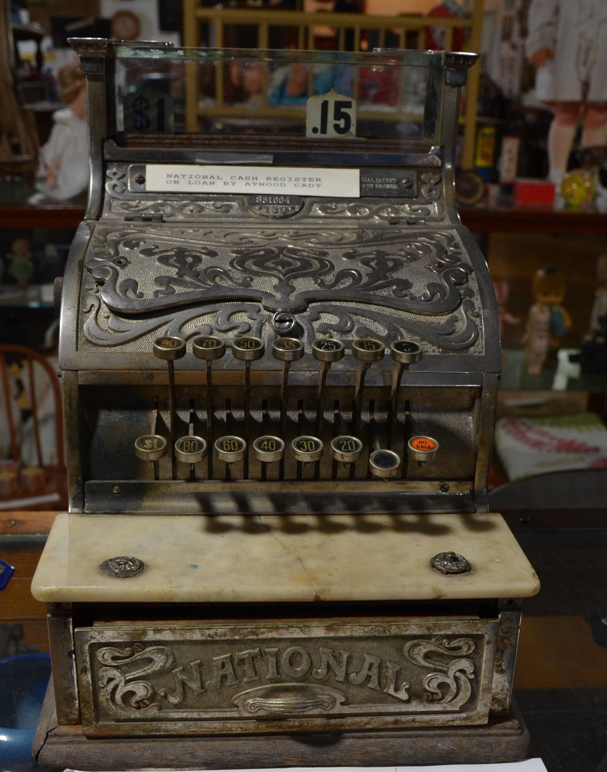 An ornate brass National cash register from 1913 on a wooden counter in a small museum, its number keys and drawer facing the camera