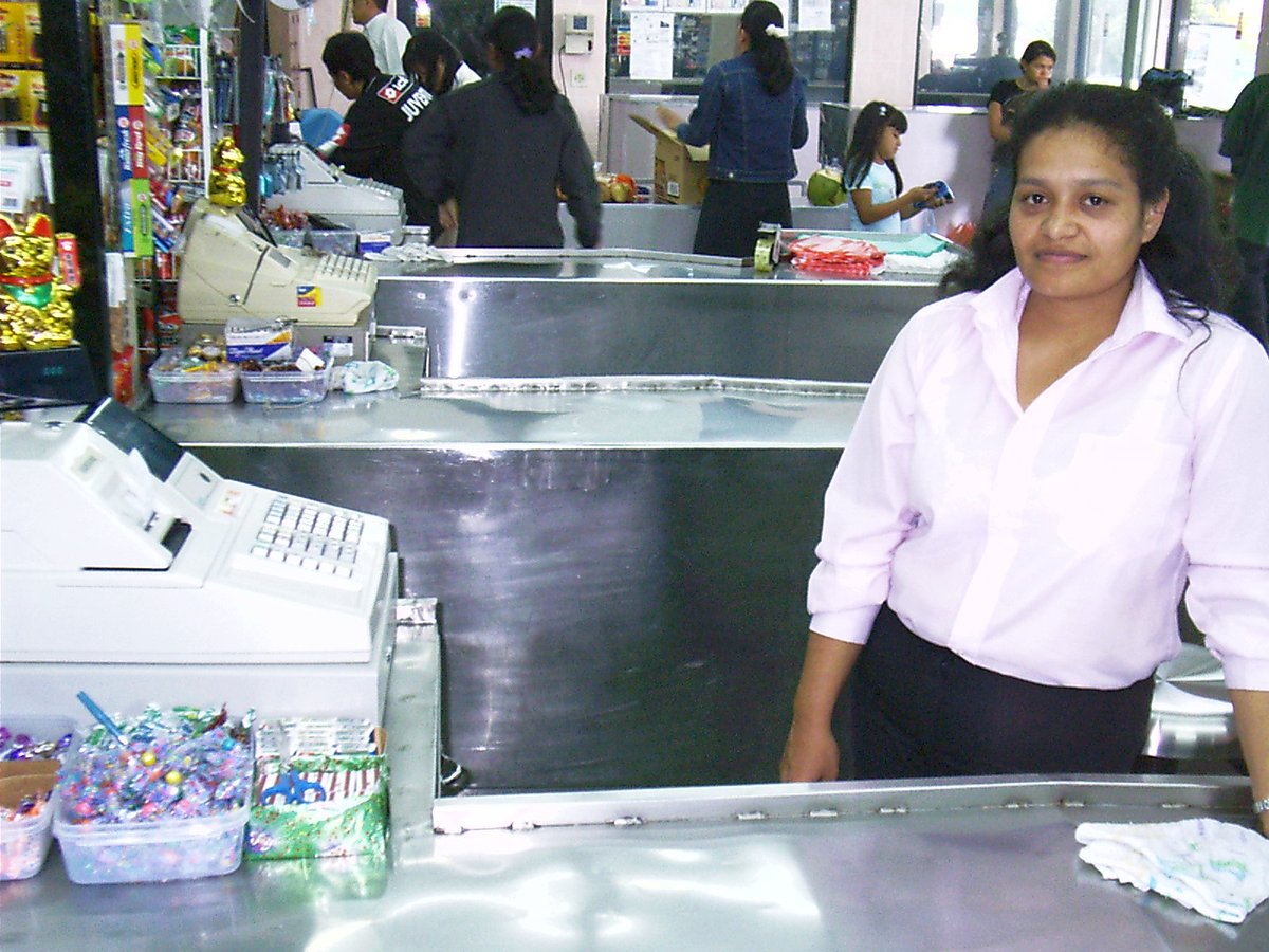 A young woman in a pink shirt stands smiling behind the counter of a grocery store beside an old electronic cash register