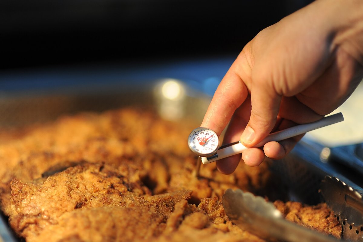 A food-service worker holding a dial-face probe thermometer in a tray of cooked food on a serving line