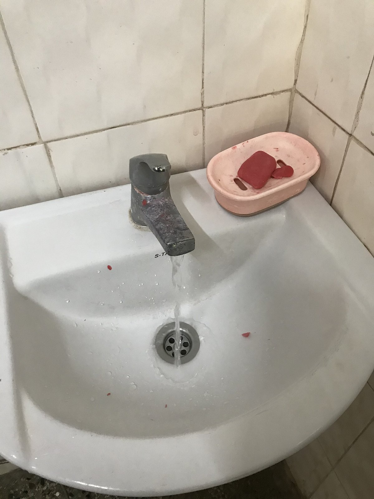 A white basin with a running tap and a pink dish of soap on its rim, against white tiles