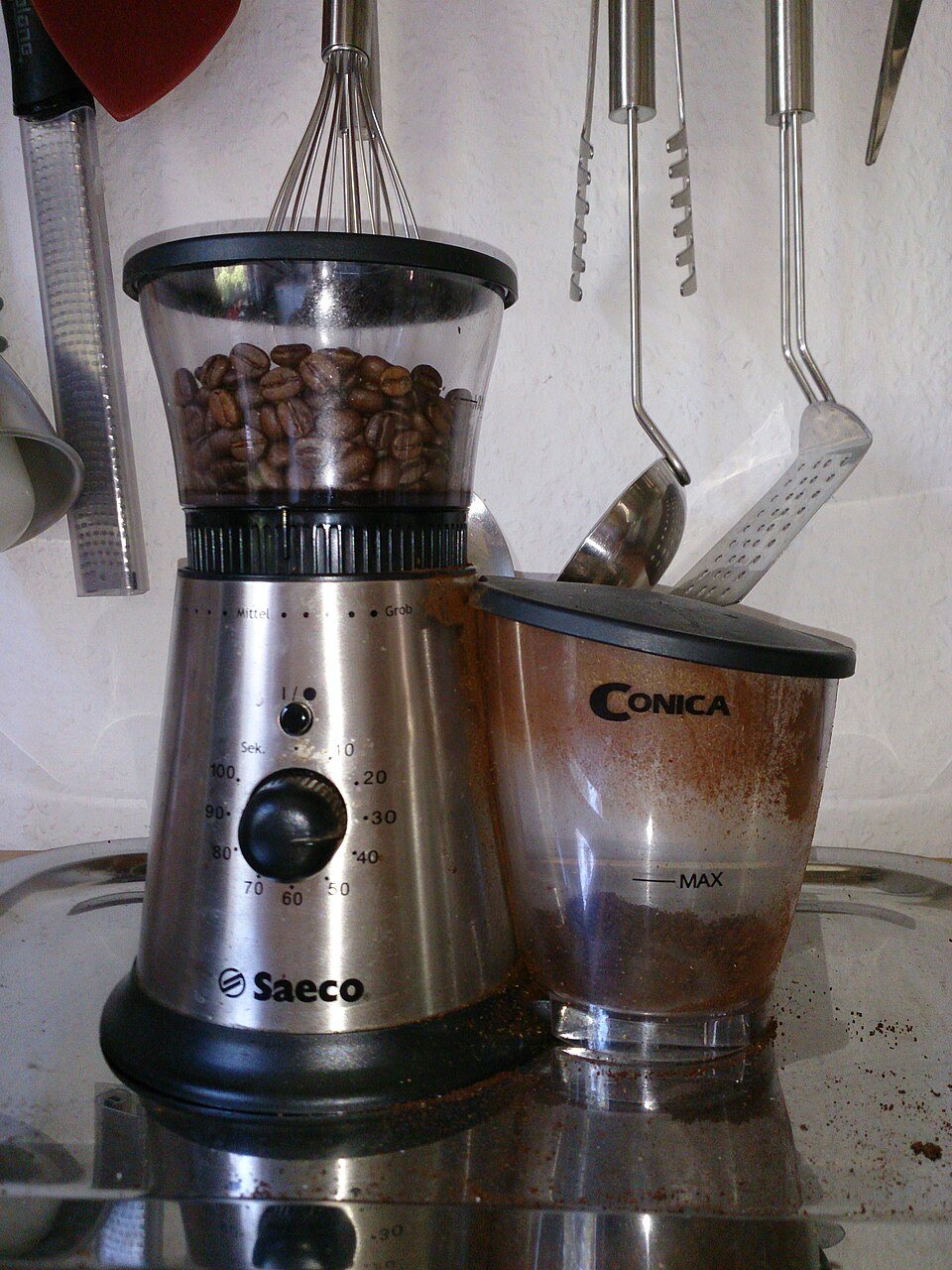 A steel home burr grinder with beans in its hopper and a container of ground coffee beside it, on a kitchen tray