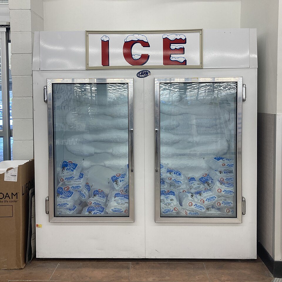 A white freezer cabinet with glass doors and a large ICE sign, stacked inside with bags of ice