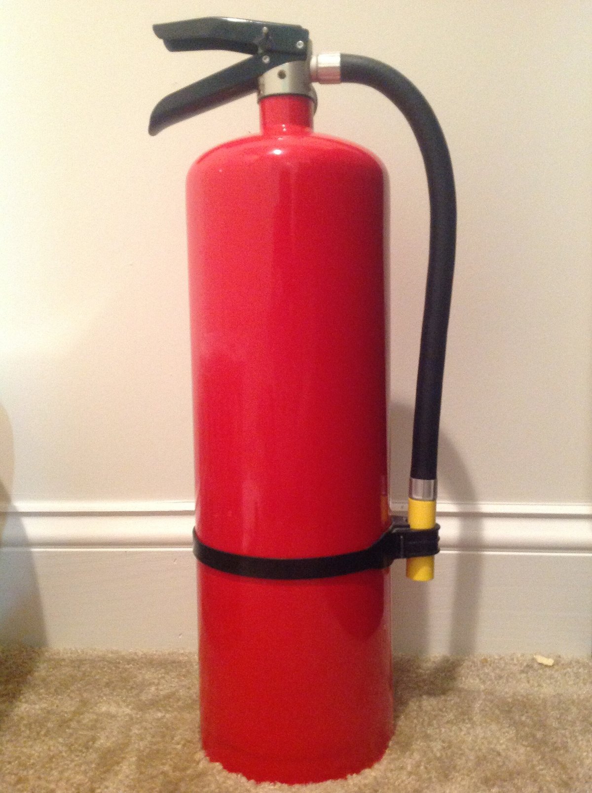 A red all-purpose fire extinguisher standing on a carpeted floor against a white wall