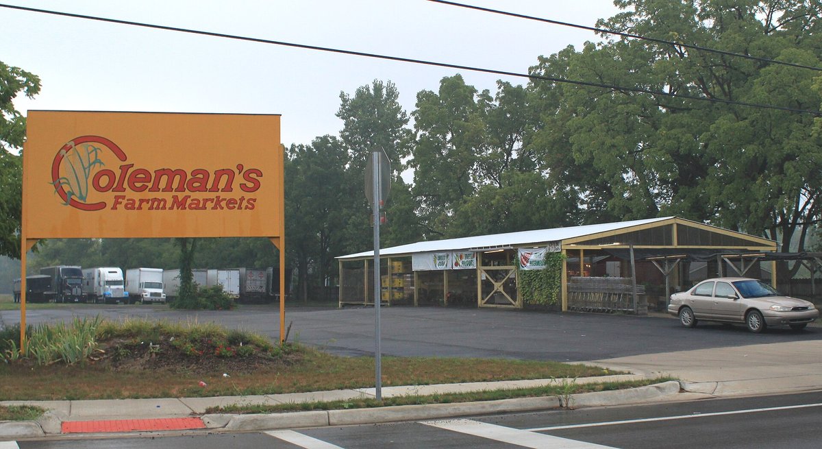 A roadside farm market building with an orange Coleman's Farm Markets sign, open-sided produce shed and a car parked in front