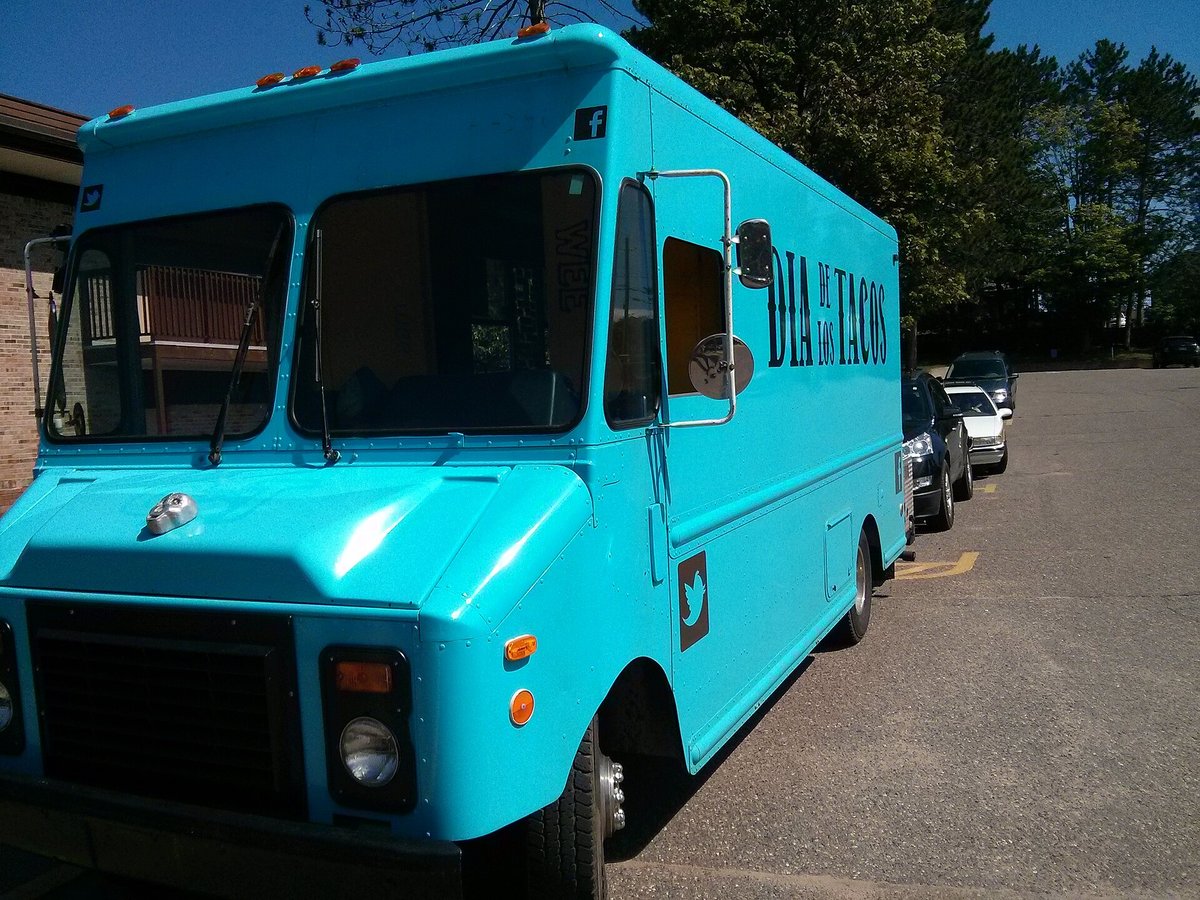 A bright turquoise step-van food truck parked in a lot under trees, its service window shut