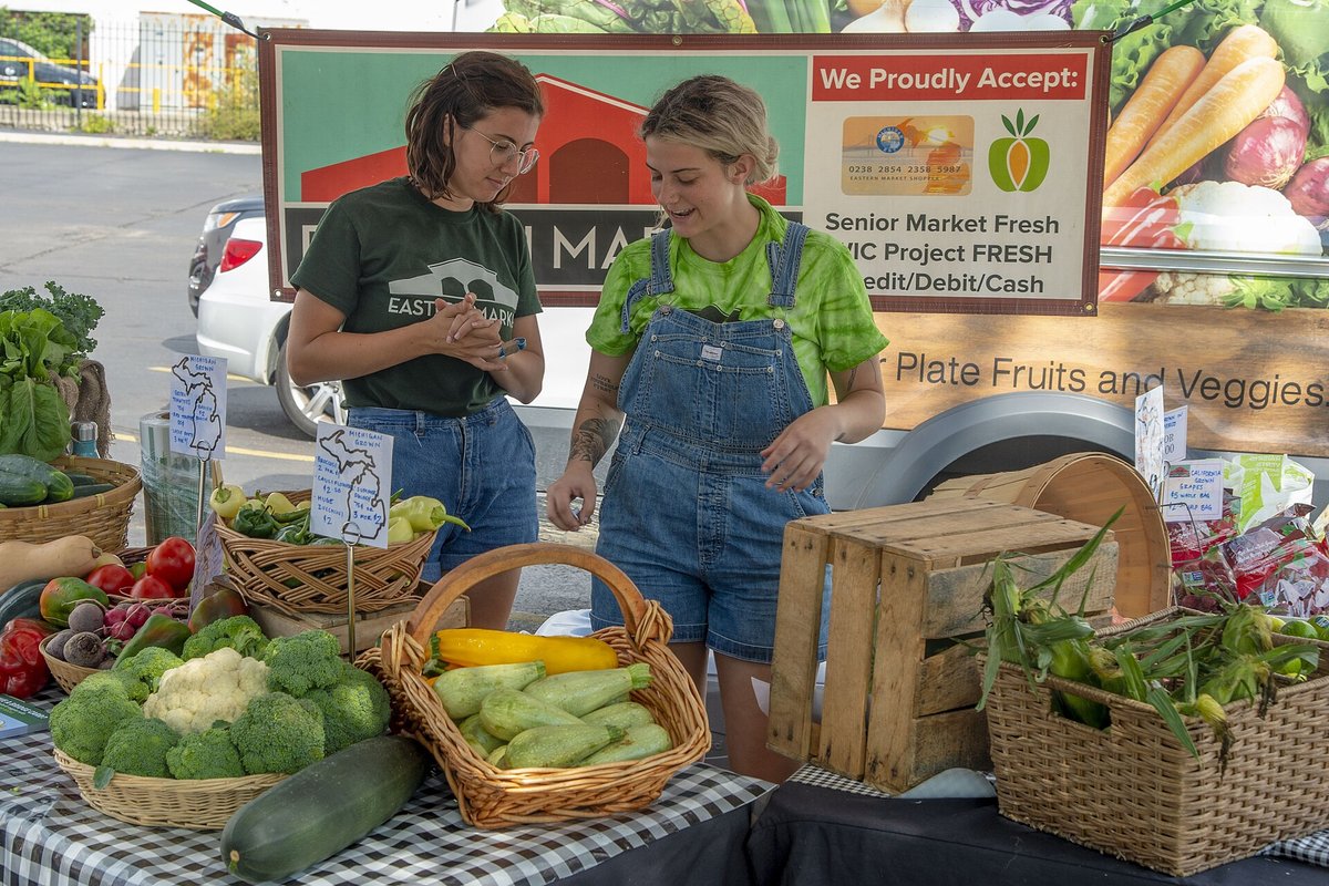 Two vendors in green shirts behind a stall of baskets of tomatoes, squash and greens, one of them in denim overalls, with a sign listing the food-assistance programmes the stand accepts