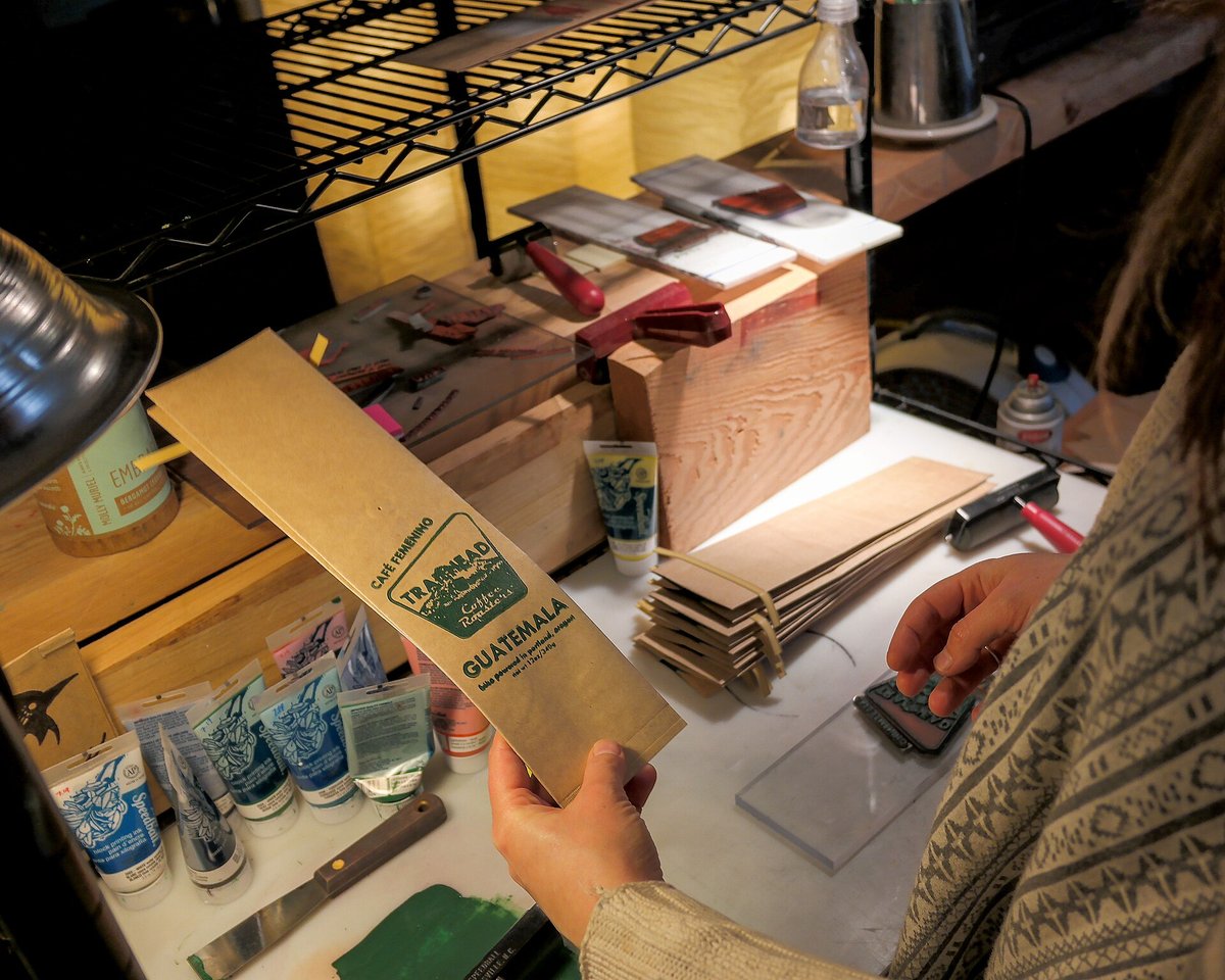 A person at a workbench presses a hand-cut printing block onto a stack of kraft coffee bags; a finished bag with a green printed logo is held up beside the stack