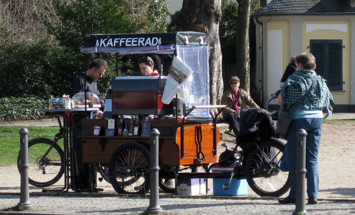 A cargo bicycle fitted as a coffee bar under a canopy reading Kaffeerad, with an espresso machine, bottles on the shelf and a cool box underneath, parked on a park path