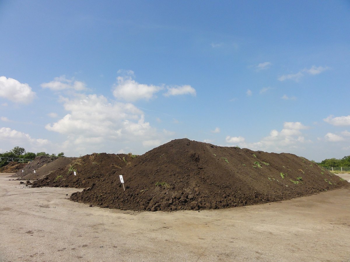 A long low pile of dark compost on a bare concrete-and-gravel yard under a blue sky with white clouds