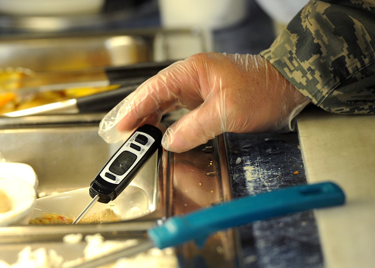 A gloved hand pressing a black digital probe thermometer into a steam-table pan of food on a serving line