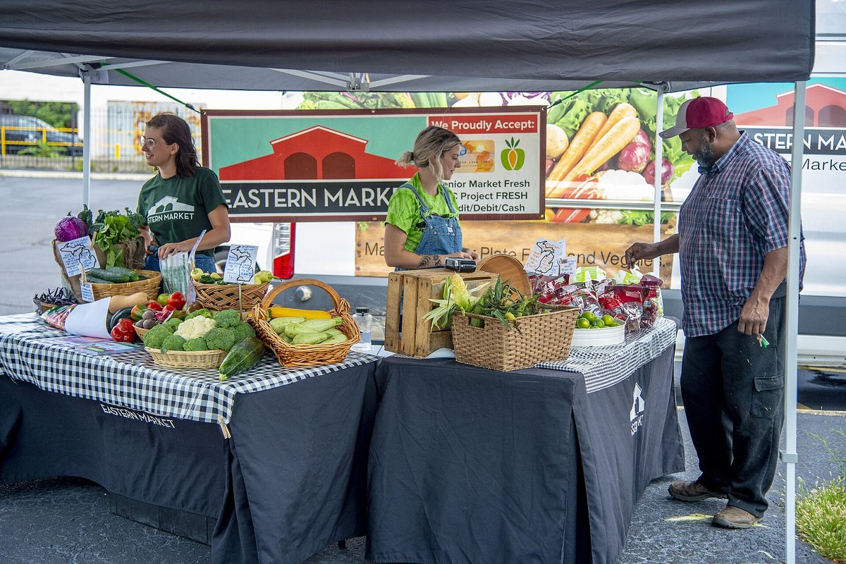 Two vendors under a black canopy at a farmers-market stand with baskets of vegetables on checked tablecloths, a customer in a red cap leaning in to look, and an Eastern Market banner behind