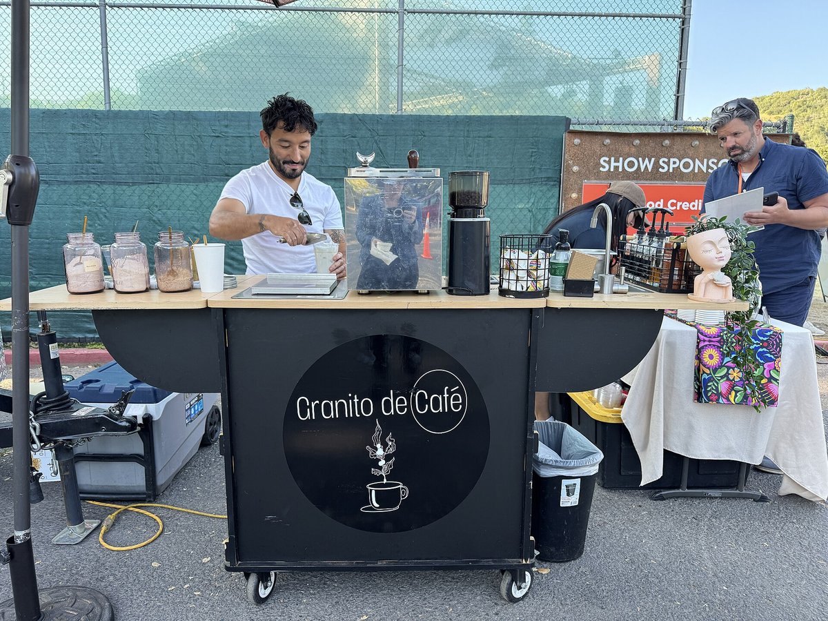 A black mobile coffee cart with a round white logo on its front, a barista behind it with jars, a grinder and brewing gear on top, set up outdoors at an evening event