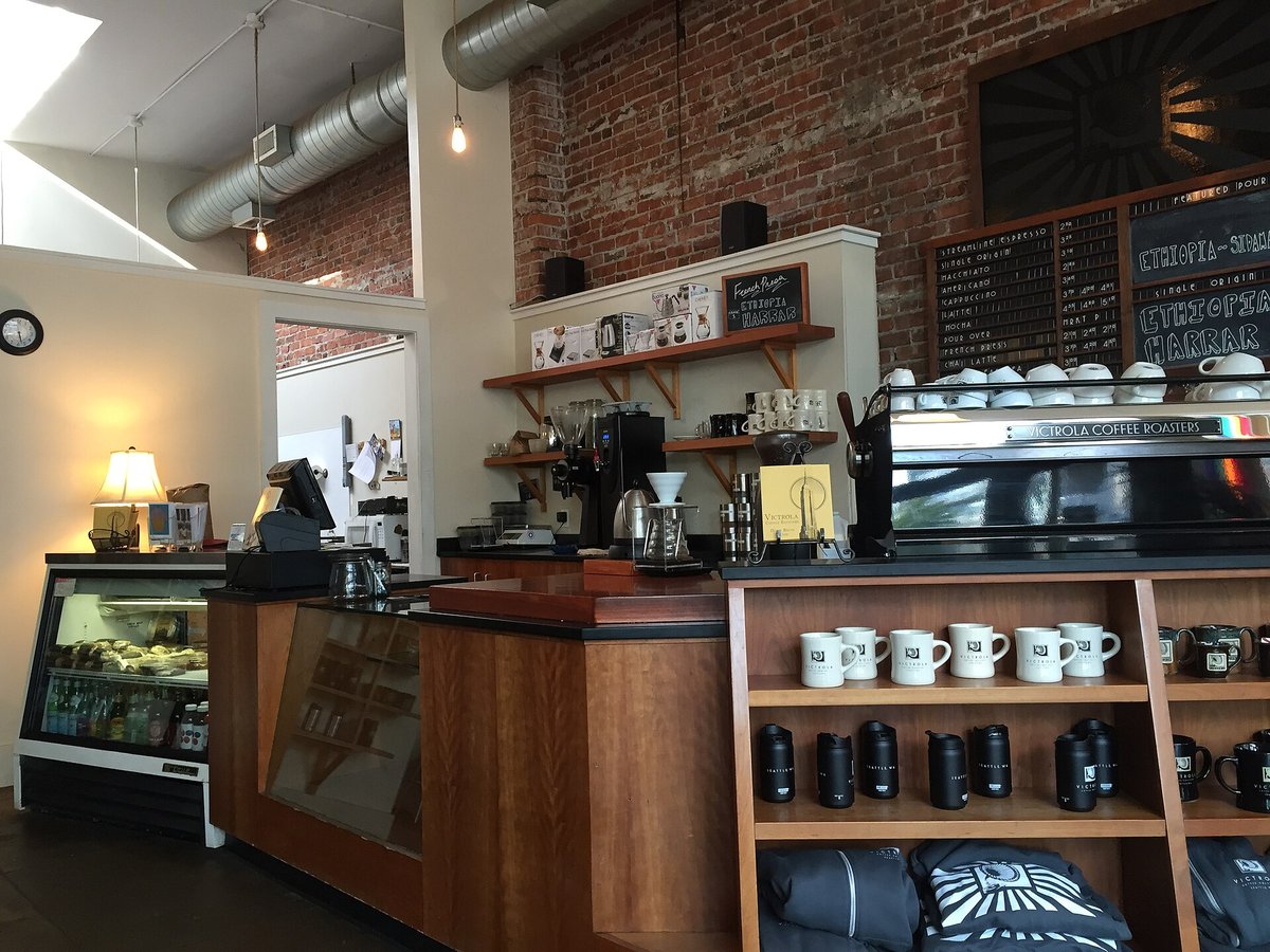 The counter of a Seattle roastery café: a pastry case on the left, the register and an espresso machine behind a wooden counter, shelves of mugs and bags of coffee, a chalkboard menu on a brick wall