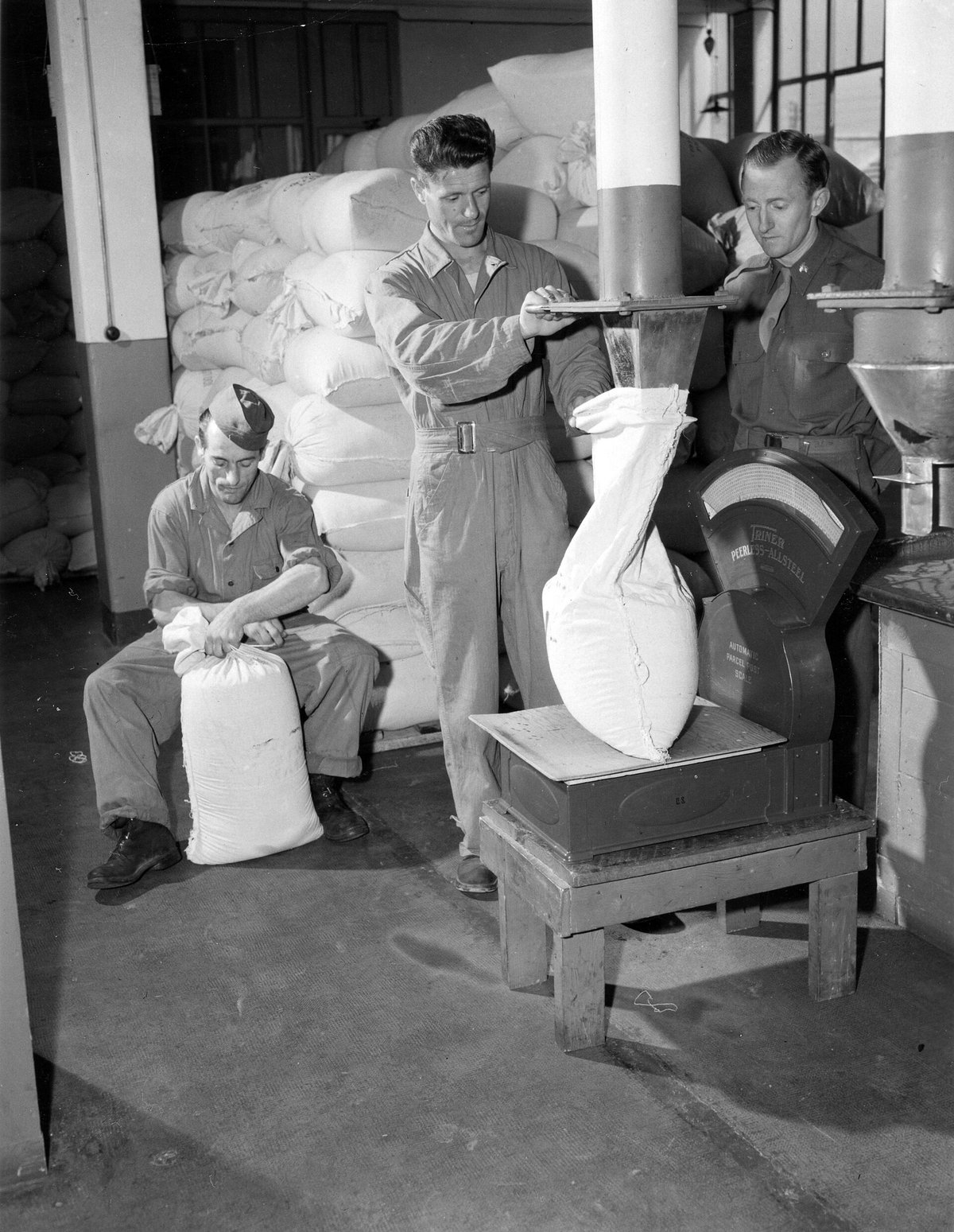 Black-and-white photograph of three soldiers in a roasting plant: one holds a white cloth sack under a steel spout so coffee drops into it while it sits on a large parcel scale; a second ties a filled sack; stacks of full sacks behind
