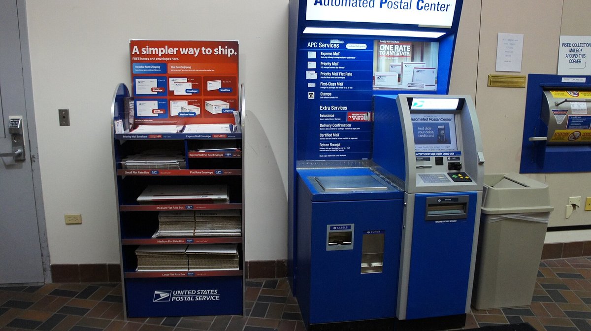 A post office lobby with a self-service postage kiosk and a red, white and blue rack of Priority Mail flat-rate boxes and envelopes