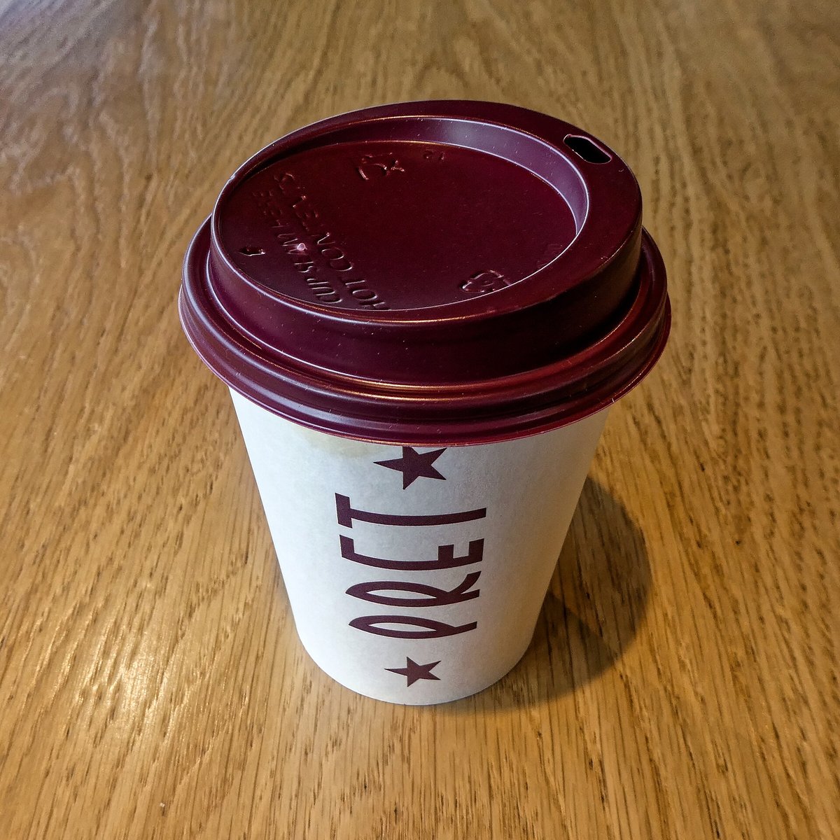 A white paper takeaway cup with a burgundy plastic sip lid pressed on, standing on a wooden table, the word Pret printed down the side