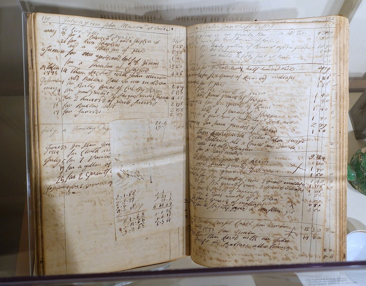 An open eighteenth-century account book in a museum case, both pages filled with columns of handwritten entries and sums in brown ink