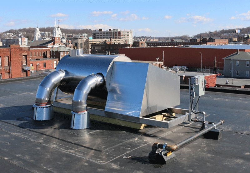 A stainless-steel afterburner with two curved inlet pipes sitting on a flat roof, with brick buildings and church steeples of a city behind it