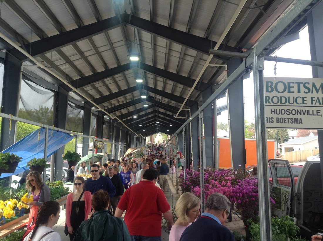 A crowded covered farmers market, shoppers walking between stalls of flowers and produce